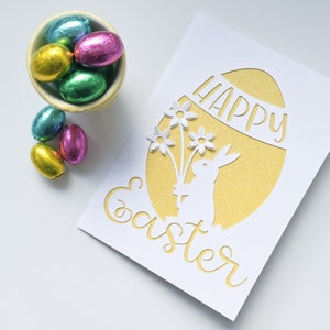 Puede incluir: Una tarjeta blanca con un huevo de purpurina dorada recortado. El huevo tiene las palabras "Happy Easter" escritas en &eacute;l. Un conejito blanco sosteniendo flores est&aacute; recortado dentro del huevo. Hay huevos de Pascua de chocolate en un taz&oacute;n a la izquierda de la tarjeta.