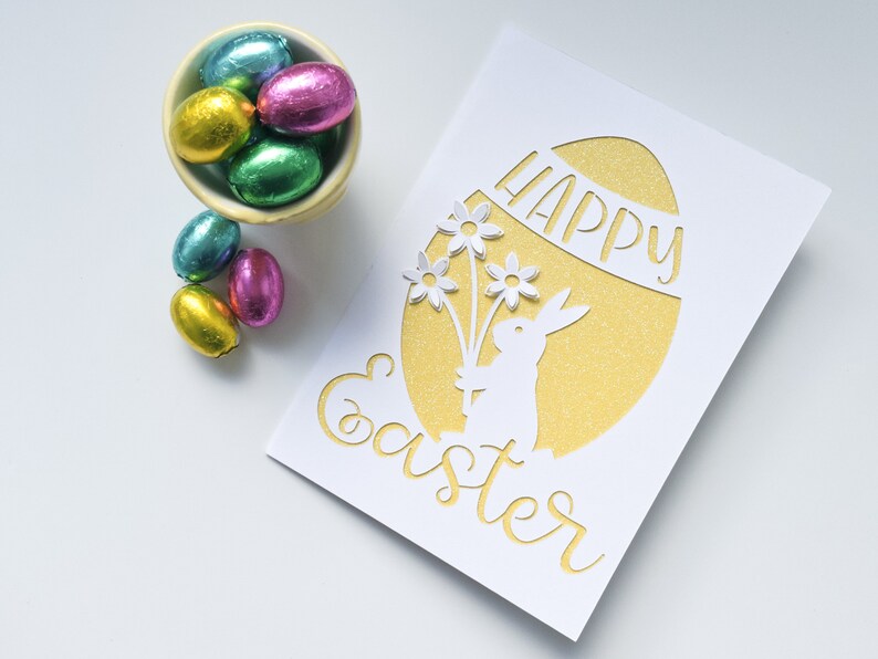 Puede incluir: Una tarjeta blanca con un huevo de purpurina dorada recortado. El huevo tiene las palabras "Happy Easter" escritas en &eacute;l. Un conejito blanco sosteniendo flores est&aacute; recortado dentro del huevo. Hay huevos de Pascua de chocolate en un taz&oacute;n a la izquierda de la tarjeta.