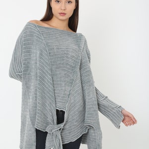 May include: A grey knitted sweater with a loose fit and a knotted detail at the front. The sweater has a slightly off-the-shoulder neckline and long sleeves.