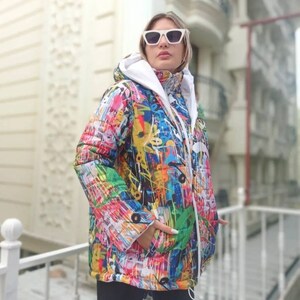 May include: A colorful, hooded jacket with a graffiti-style design. The jacket features a vibrant mix of colors including blue, yellow, red, and green. It has a white interior and a drawstring. The person is wearing white sunglasses.