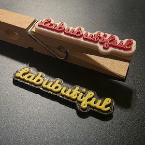Può includere: Una molletta di legno con un design "Labubutiful" rosso e bianco attaccato. È visibile anche un design "Labubutiful" giallo e nero separato. Gli oggetti sono su una superficie scura.