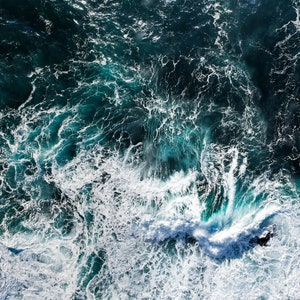 Puede incluir: Vista aérea de un océano azul oscuro con olas blancas espumosas que rompen contra la costa rocosa.