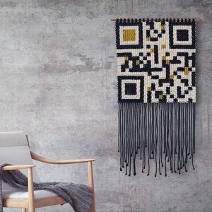 May include: A white armchair with a gray throw blanket sits in front of a gray concrete wall. A black and white woven wall hanging with gold accents hangs on the wall. The wall hanging is a QR code design.
