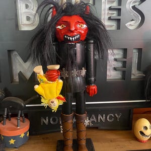 May include: A wooden nutcracker figure with a red face, sharp white teeth, and long black hair, featuring two horns on its head. It wears a black outfit with silver snowflake accents and brown boots, while holding a small doll upside down.