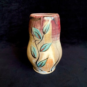 以下が含まれることがあります： 青、赤、茶色の色合いでスタイリッシュな葉が手描きされた、ユニークな有機的な形をしたセラミックの花瓶。花瓶はテクスチャのある釉薬がかけられた表面と、わずかに不規則な縁を持っています。