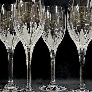 May include: Four clear crystal wine glasses with a leaf design etched into the bowl. The glasses have long stems and round bases. The glasses are arranged in a row against a dark background. The glasses are elegant and suitable for formal occasions.