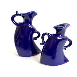 May include: Two cobalt blue ceramic pitchers with a glossy finish. Each pitcher has a curved handle and a unique spout design. The pitchers are displayed against a white background, highlighting their shape and color.