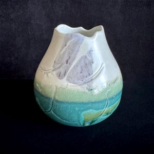 May include: A handmade ceramic vase with an unusual, organic form. The vase has a white top, fading into a lavender and green midsection, and a turquoise base. The design incorporates leaf-like patterns, enhancing its artistic quality.