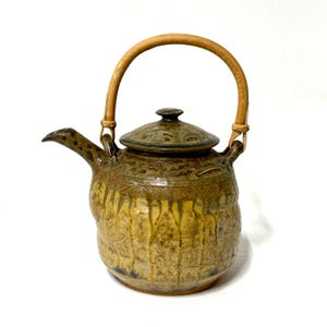 May include: A ceramic teapot with a bamboo handle and lid. The teapot has a textured, glazed surface in shades of brown and yellow. The lid is a darker green with a floral pattern. The handle is made of curved bamboo.