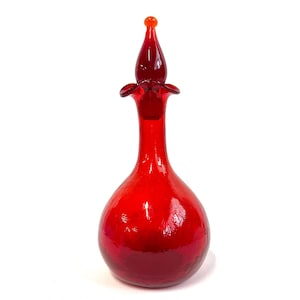May include: A vibrant red glass bottle with a bulbous body and a long, slender neck. The bottle is topped with a decorative stopper featuring a teardrop shape and a small orange orb. The glass has a glossy finish.
