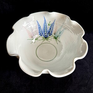 May include: White ceramic bowl with a scalloped edge. The bowl features a hand-painted floral design with blue, pink, and green accents. A green circle is painted in the center of the bowl. The bowl is likely used for serving or display.