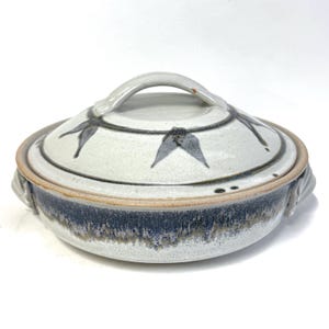 May include: A ceramic lidded serving dish with a light blue and white glaze. The lid features a handle and a leaf design in dark blue. The dish has a dark blue band around the base and two handles on the sides.