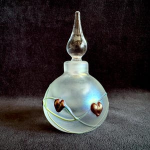 May include: A round, frosted glass perfume bottle with an iridescent finish. The bottle features a clear, teardrop-shaped glass stopper and is decorated with thin, light green glass vines and small, dark reddish-brown heart-shaped glass accents.