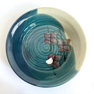 May include: A ceramic plate in teal and cream. The plate has a spiral design in the centre and a painted floral pattern. A sticker on the bottom reads "Pottery Gallery Studio". The plate is suitable for serving food.