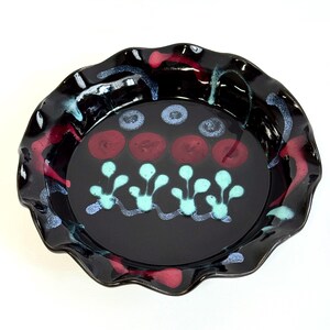 May include: A black ceramic bowl with a ruffled edge. The bowl's interior features a design of red and blue circles, and turquoise plant-like shapes. Red and blue paint accents the rim.