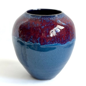 May include: A handcrafted ceramic vase with a rounded shape. The upper portion is a deep burgundy color with a speckled texture, transitioning to a smooth, glossy blue base. The interior is dark. A decorative piece for the home.
