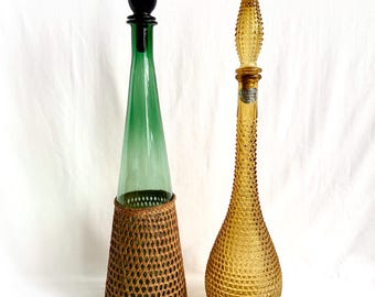May include: Two vintage glass decanters with unique stoppers. The decanter on the left is green glass with a black stopper and a woven brown wicker base. The decanter on the right is amber glass with a clear glass stopper and a textured surface.