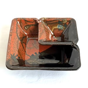 May include: A two-tiered ceramic dish with a square shape and a distinctive design. The top tier is smaller than the bottom tier. The dish has a red and black colour scheme with abstract patterns. The edges are dark brown, and the overall aesthetic is artistic and handcrafted.