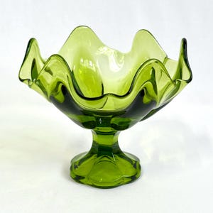 May include: A translucent olive green glass bowl featuring a wide, ruffled, and scalloped rim. The bowl is supported by a sturdy, multi-faceted pedestal base that flares out at the bottom, creating a distinct footed silhouette for this glass vessel.