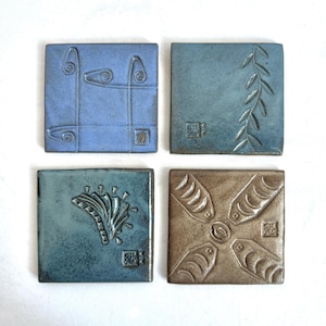 May include: Four square ceramic tiles with different designs. The tiles are in shades of blue, brown, and beige. The designs include a stylized plant, a geometric pattern, a stylized flower, and a stylized cross.