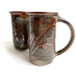 May include: Two tall ceramic mugs featuring a dark gray and brown mottled glaze with a hand-painted botanical pattern of reddish-brown pussy willow branches. Each mug has a cylindrical body, a wide rim, and a curved handle on the side.