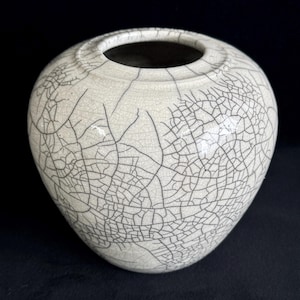 May include: A round ceramic vase with a wide, circular opening at the top. The exterior surface is off-white and features a prominent, dark black crackle glaze pattern that covers the entire body, creating a dense network of fine lines.