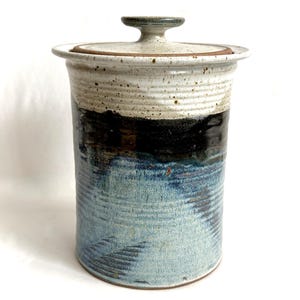 May include: A cylindrical ceramic canister with a matching lid featuring a knob handle. The upper portion is a speckled off-white, transitioning into a deep black band, and finishing with a textured, light blue base with subtle horizontal ridges.