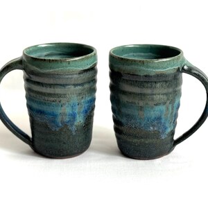 Puede incluir: Dos tazas de cerámica hechas a mano con un diseño texturizado y acanalado. Las tazas presentan un degradado de colores, que va desde un borde turquesa hasta tonos de azul y verde oscuro. Cada taza tiene un asa curva.