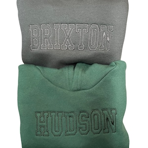 May include: Two hooded sweatshirts stacked on top of each other. The top sweatshirt is gray with the word "BRIXTON" embroidered in white. The bottom sweatshirt is green with the word "HUDSON" embroidered in black.