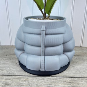 May include: A grey ceramic planter shaped like a puffer jacket with a black base. The planter has a zipper detail and is filled with a small plant.