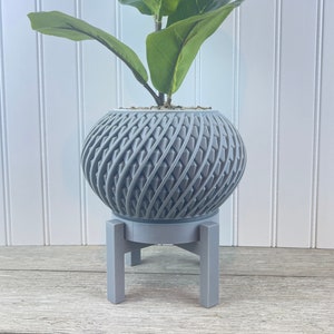May include: A gray, spherical planter with a textured, woven design, holding a green plant. The planter sits on a matching gray stand with a three-legged base. The background features white vertical paneling and a wooden surface.