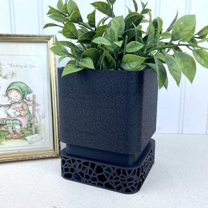 May include: A black planter with a geometric pattern base, holding a green leafy plant. The planter is made of a textured material.