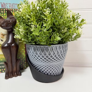 May include: A gray, patterned planter with a black base holds a vibrant green plant with small white flowers. A dark brown cat statue and a book are visible in the background. The planter has a woven design.