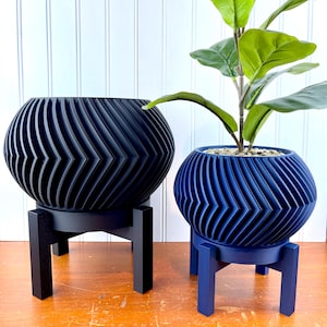Op de afbeelding: Twee decoratieve plantenbakken, één zwart en één blauw, elk met een chevron-ontwerp. De plantenbakken staan op bijpassende standaards. De blauwe plantenbak bevat een groene plant.