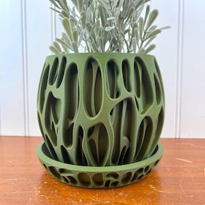 Pode incluir: Um vaso verde, impresso em 3D, com um design orgânico aberto. O vaso assenta sobre um pires a condizer e contém uma planta falsa com folhas verde-prateadas. Um artigo decorativo para decoração de casa.