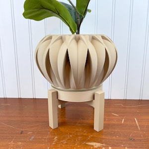 May include: A beige ceramic planter with a unique ribbed design sits on a matching beige stand with four legs. The planter is empty and ready for a plant.