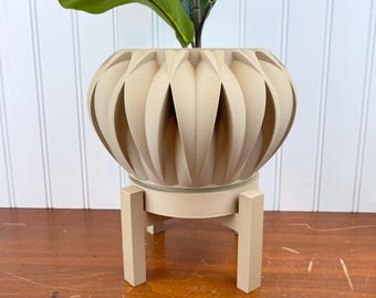 May include: A beige ceramic planter with a unique ribbed design sits on a matching beige stand with four legs. The planter is empty and ready for a plant.