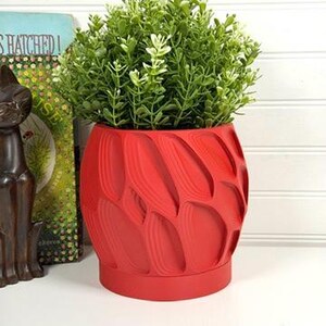 Puede incluir: Una maceta roja vibrante con un patrón texturizado en forma de hoja. La maceta está llena de una exuberante planta verde. Una estatua decorativa de gato y un libro con el texto "Hatched!" están en el fondo.