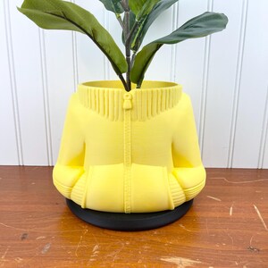 Pode incluir: Um vaso de cerâmica amarelo em forma de suéter com zíper. O vaso tem uma base preta e contém uma planta verde.