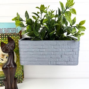 May include: A rectangular wall-mounted planter with a textured grey brick pattern on the exterior, containing a lush green artificial plant with pointed leaves. The planter is positioned next to a dark brown wooden cat figurine and a book.