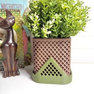 May include: A brown and green decorative planter with a lattice design, holding a vibrant green plant with white accents. The planter has a geometric base and a square shape. A wooden cat statue and a book are in the background.
