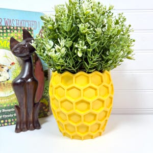 May include: A bright yellow honeycomb-patterned planter filled with green and white faux greenery. A dark brown cat figurine and a vintage children's book are in the background. The planter is a decorative item for home decor.