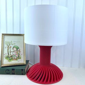 May include: A red table lamp with a white drum shade. The base of the lamp is made of a series of curved, overlapping pieces of wood.