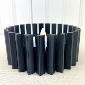 May include: Black candle holder with a circular design. The holder is made of multiple vertical slats that create a fence-like pattern.