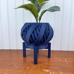 Op de afbeelding: Een marineblauwe plantenpot met een uniek geweven ontwerp staat op een bijpassende marineblauwe standaard. De pot is rond en heeft een brede opening. De standaard heeft vier poten.