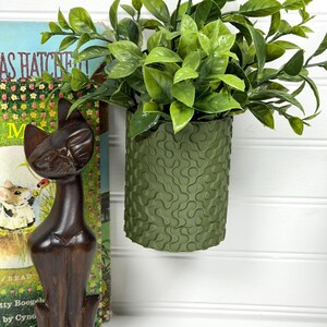 Può includere: Un vaso cilindrico verde scuro con un motivo strutturato, riempito di piante verdi e vibranti. Una statuetta di gatto in legno e un libro vintage sullo sfondo creano un'esposizione decorativa.