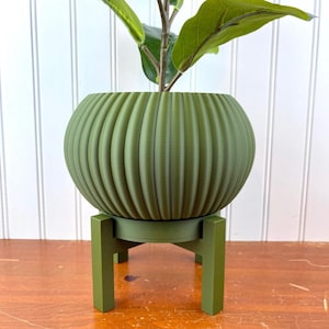 Op de afbeelding: Een groene keramische plantenpot met een geribbeld ontwerp staat op een bijpassende groene houten standaard. De pot is rond en heeft een brede opening. De standaard heeft drie poten.