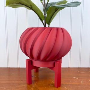 Pode incluir: Um vaso de cerâmica vermelho com um design em espiral fica em cima de um suporte de metal vermelho. O vaso está cheio de uma planta verde com folhas.