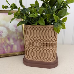 May include: A beige plant pot with a repeating oval design, containing green leafy plants. The pot rests on a dark brown base. A framed picture is visible in the background.