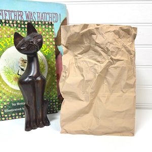 Puede incluir: Una figura de gato de madera oscura con cuello largo y orejas puntiagudas se encuentra junto a un libro infantil vintage titulado "Fletcher Was Hatched!". Una bolsa de papel marrón arrugada también está en la imagen.